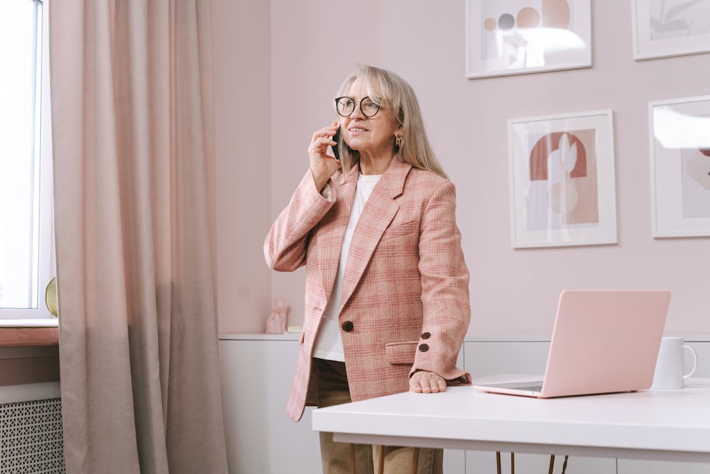 Senior woman in a pink blazer stands by a desk, talking on smartphone, in a stylish office.